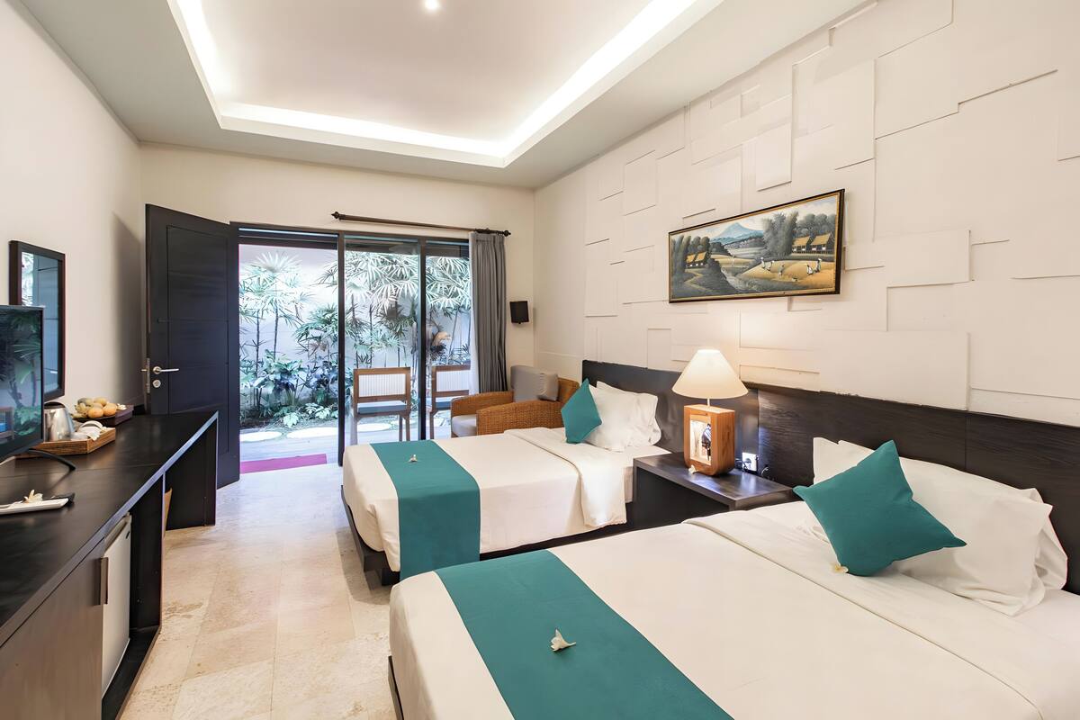 A hotel room with two beds, a desk, a TV, and a view of a garden through a sliding door.