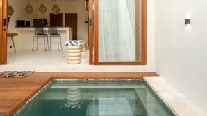 A modern indoor pool area with a wooden deck, a dining area with chairs and a table, and a kitchen in the background. - Liminal Palace Villa 2 by Betterplace (Canggu)