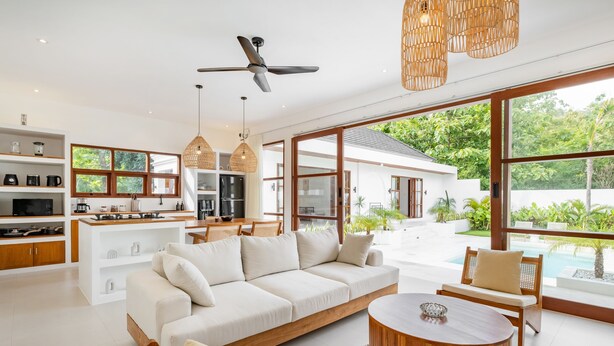Un salon moderne avec un canapé, une table basse et un coin repas. Une grande fenêtre offre une vue sur la verdure et une piscine.