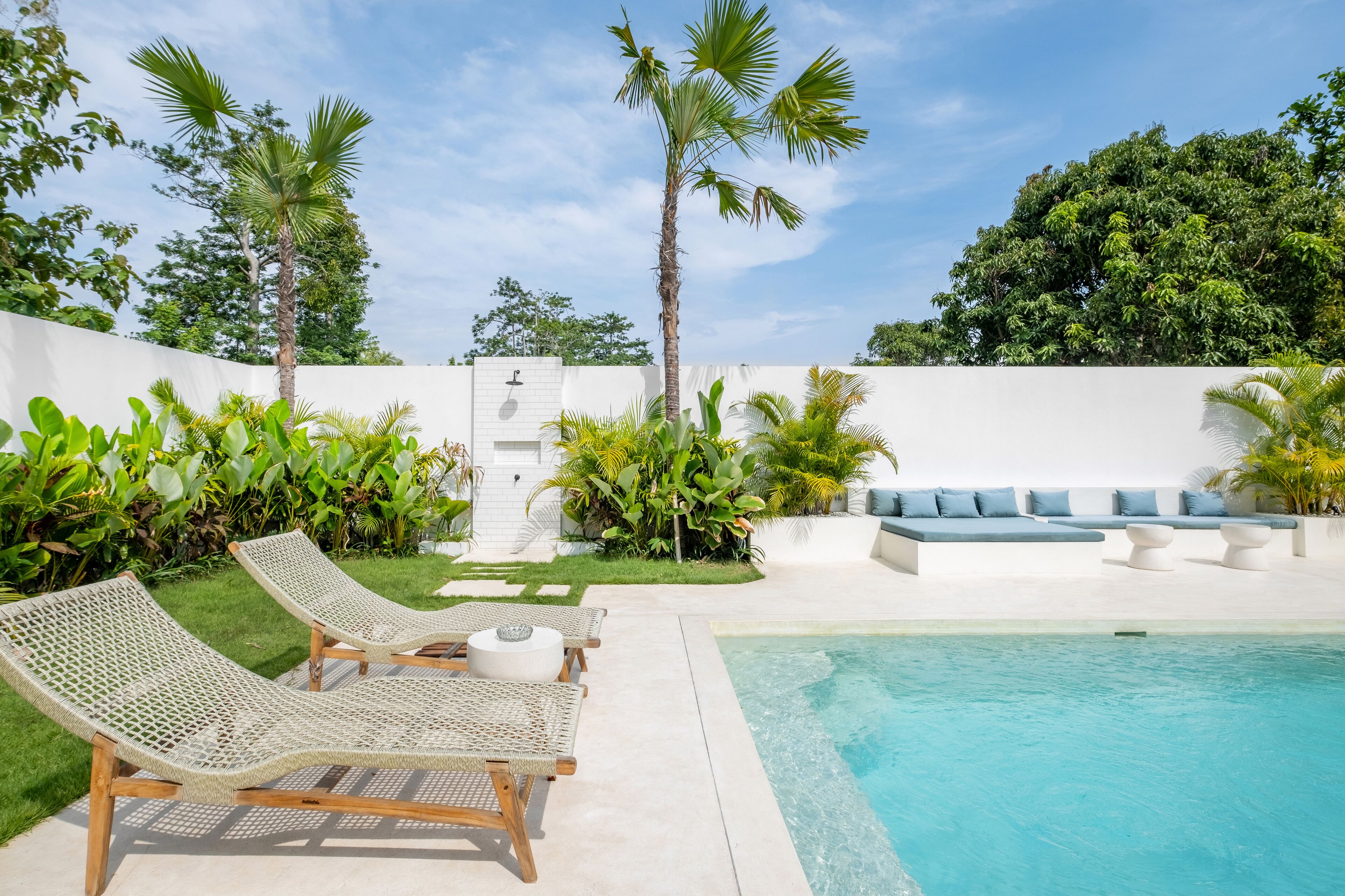 A poolside area with lounge chairs, a seating area with cushions, and lush greenery including palm trees.