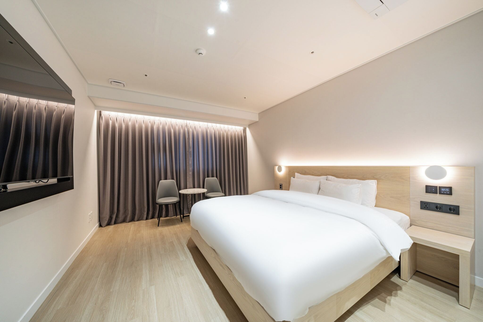 A modern hotel room with a large bed, a desk, two chairs, and a TV mounted on the wall.