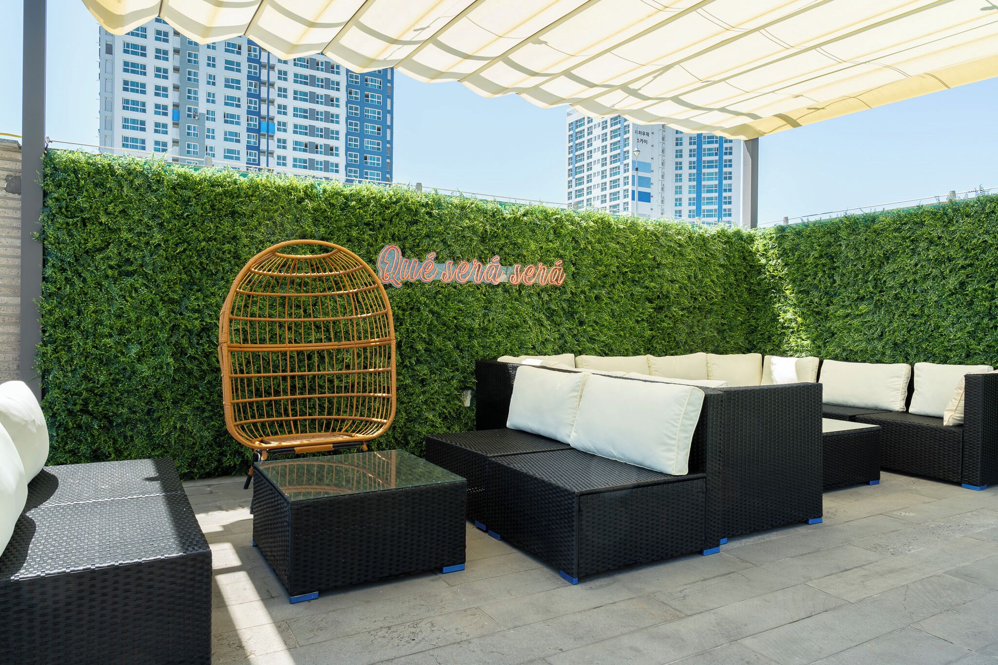 An outdoor seating area with wicker furniture and a glass table, a green hedge wall, and a cityscape in the background.