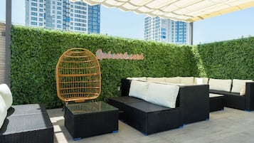An outdoor seating area with wicker furniture and a glass table, a green hedge wall, and a cityscape in the background.