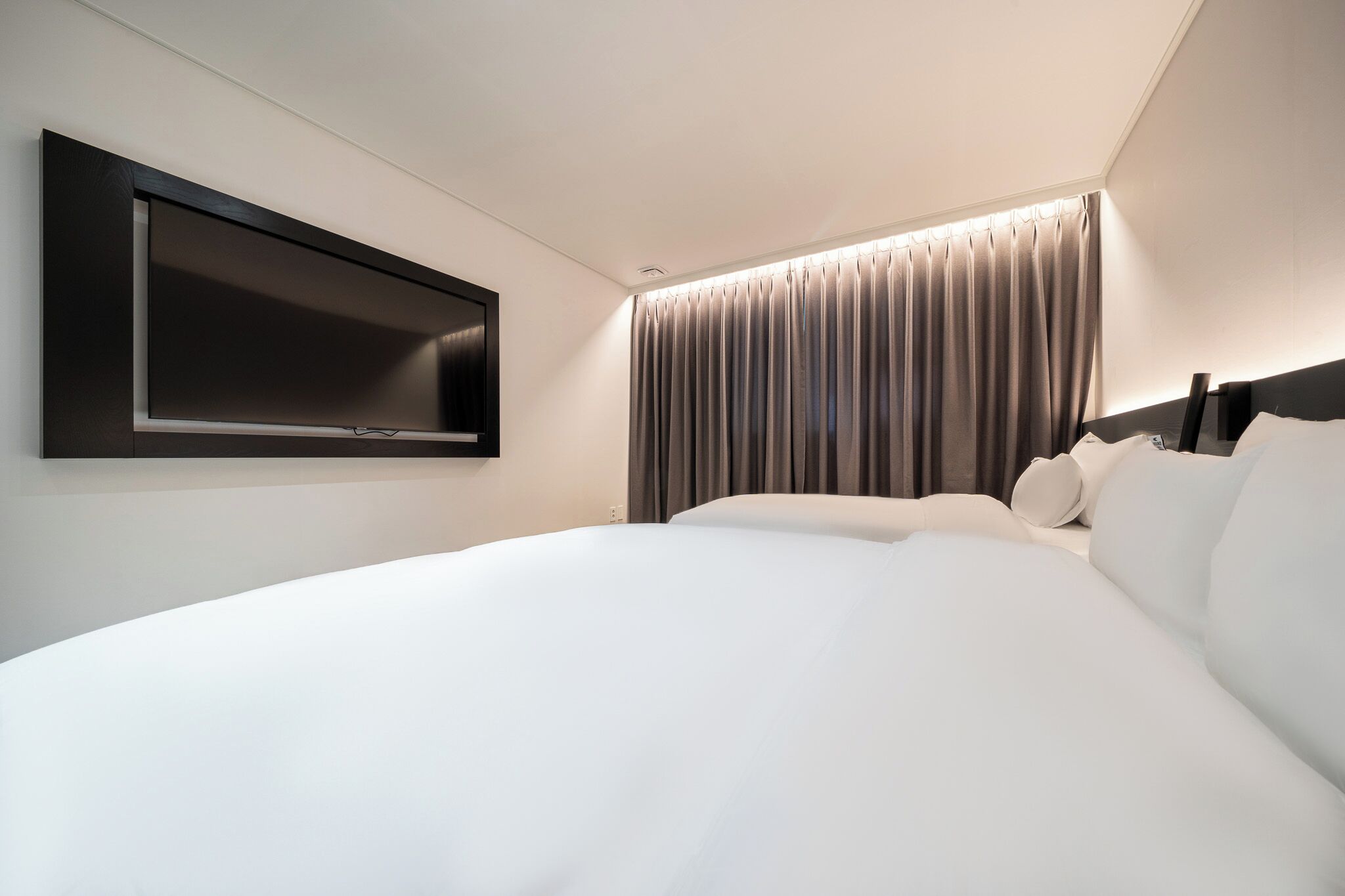 A modern hotel room with a large bed, a flat-screen TV mounted on the wall, and a window with curtains.