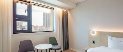 A hotel room with a bed, two chairs, a small table, a window with curtains, and a city view.