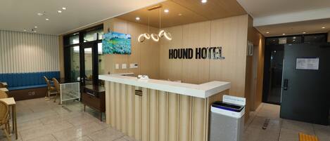 Una recepción con un cartel que dice “HOUND HOTEL” en un vestíbulo moderno, con un mostrador de madera, una mampara de vidrio y un área de estar con sillas.