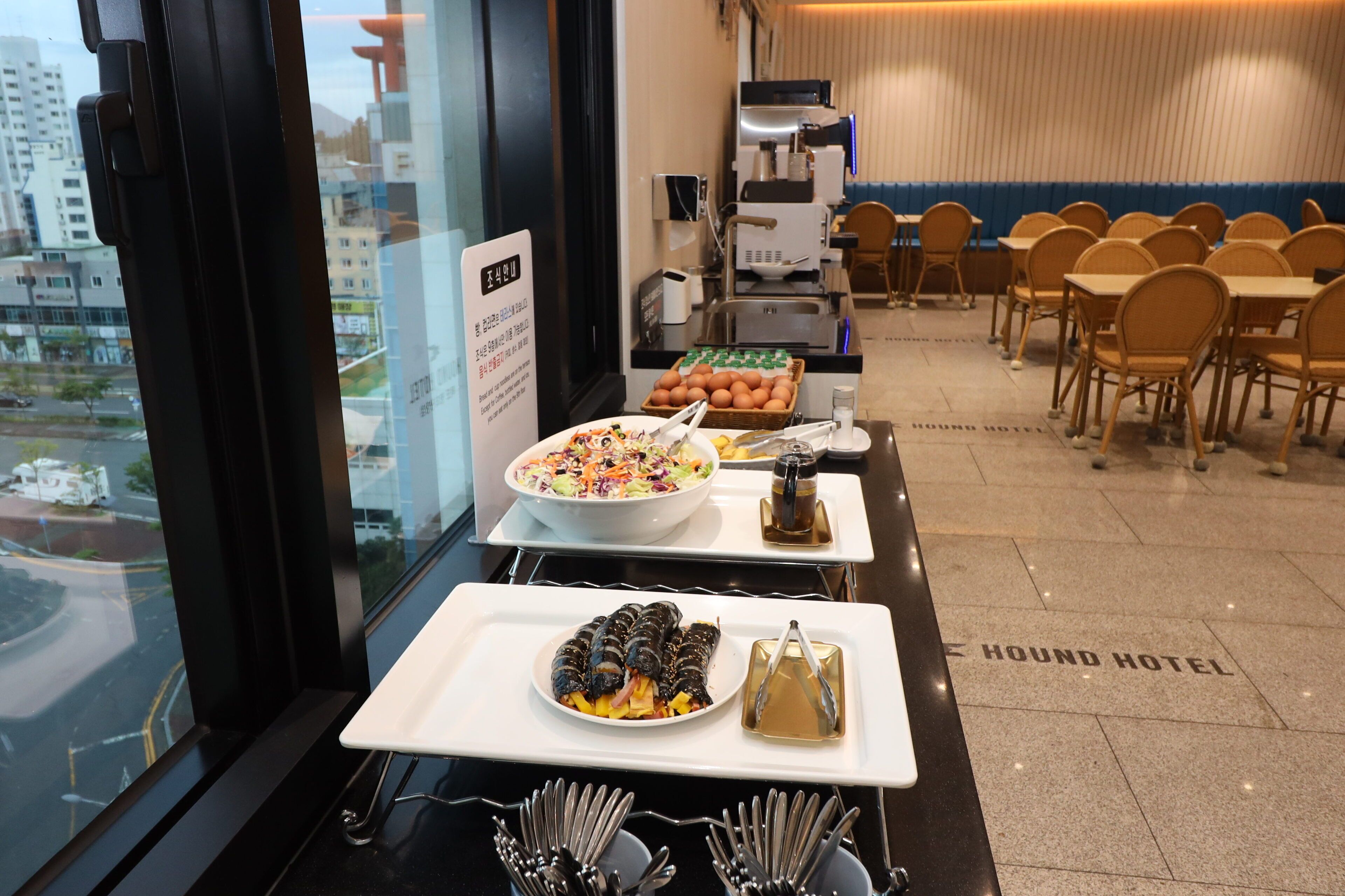 A buffet setup with a variety of dishes, including a salad, a fruit bowl, and sushi rolls, in a hotel room with a city view.
