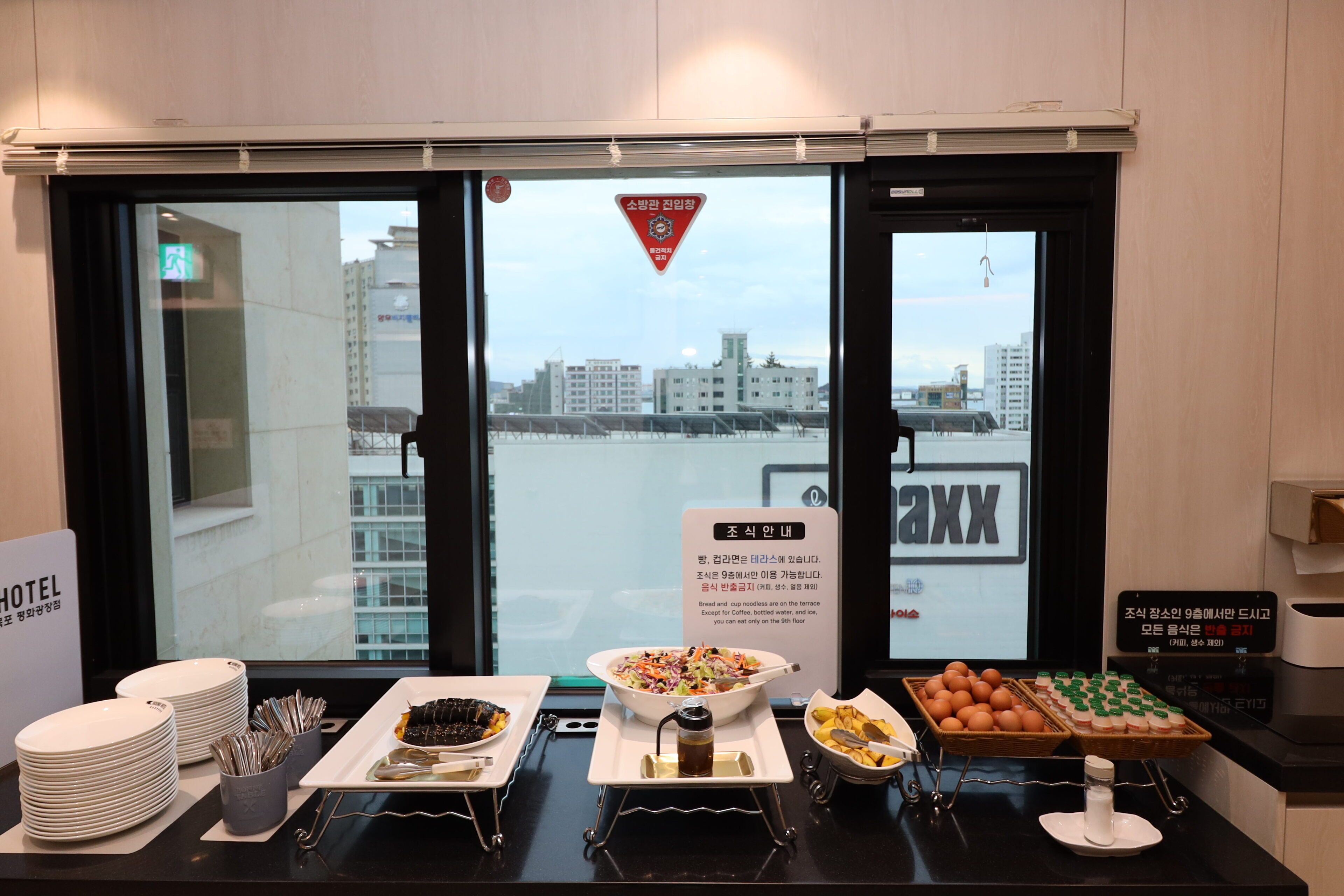 A hotel breakfast buffet with various dishes, a view of the cityscape through the window, and a sign indicating the location.