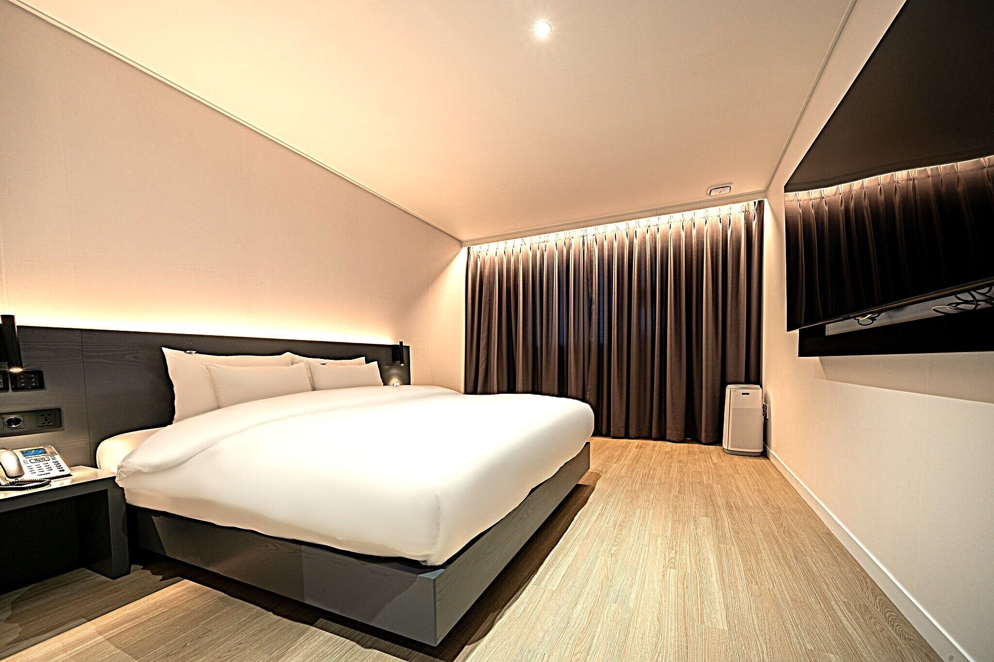 A modern hotel room with a large bed, a bedside table with a phone, and a wall-mounted TV.