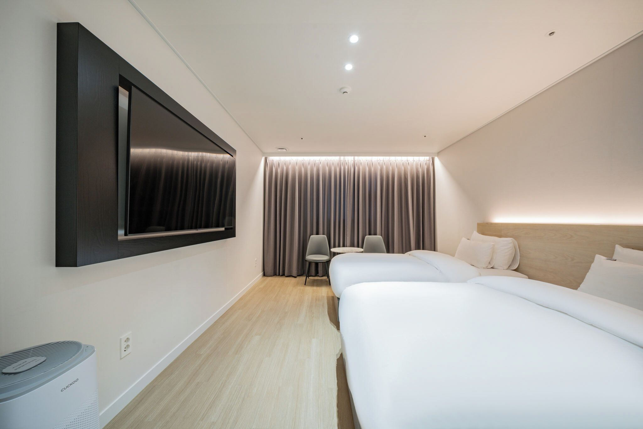 Una habitación de hotel moderna con una televisión de pantalla plana grande montada en la pared, una cama, una mesita de noche, una silla y una ventana con cortinas.