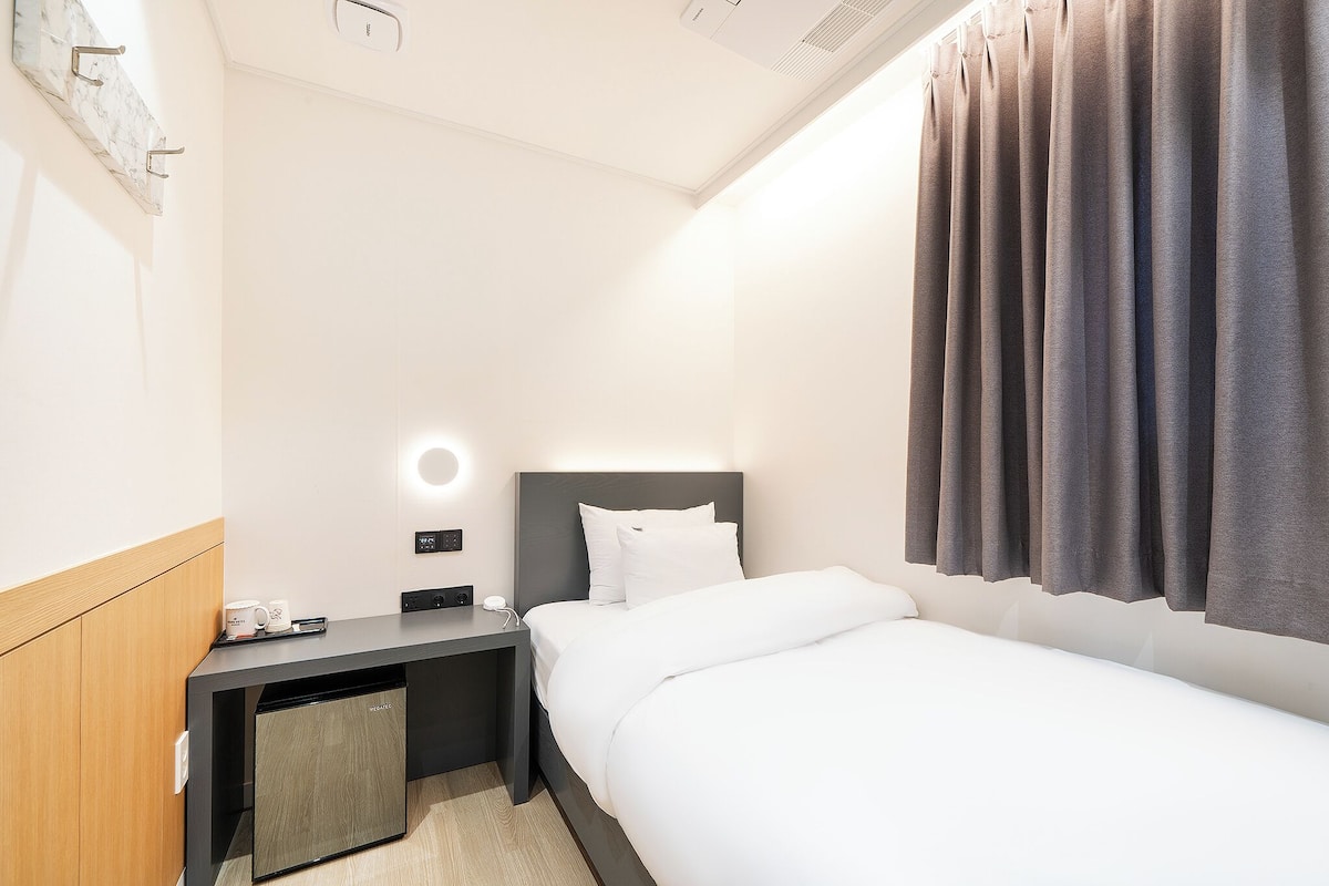 Een hotelkamer met een bed, een bureau met een lamp, een klein kastje en een wandplank.