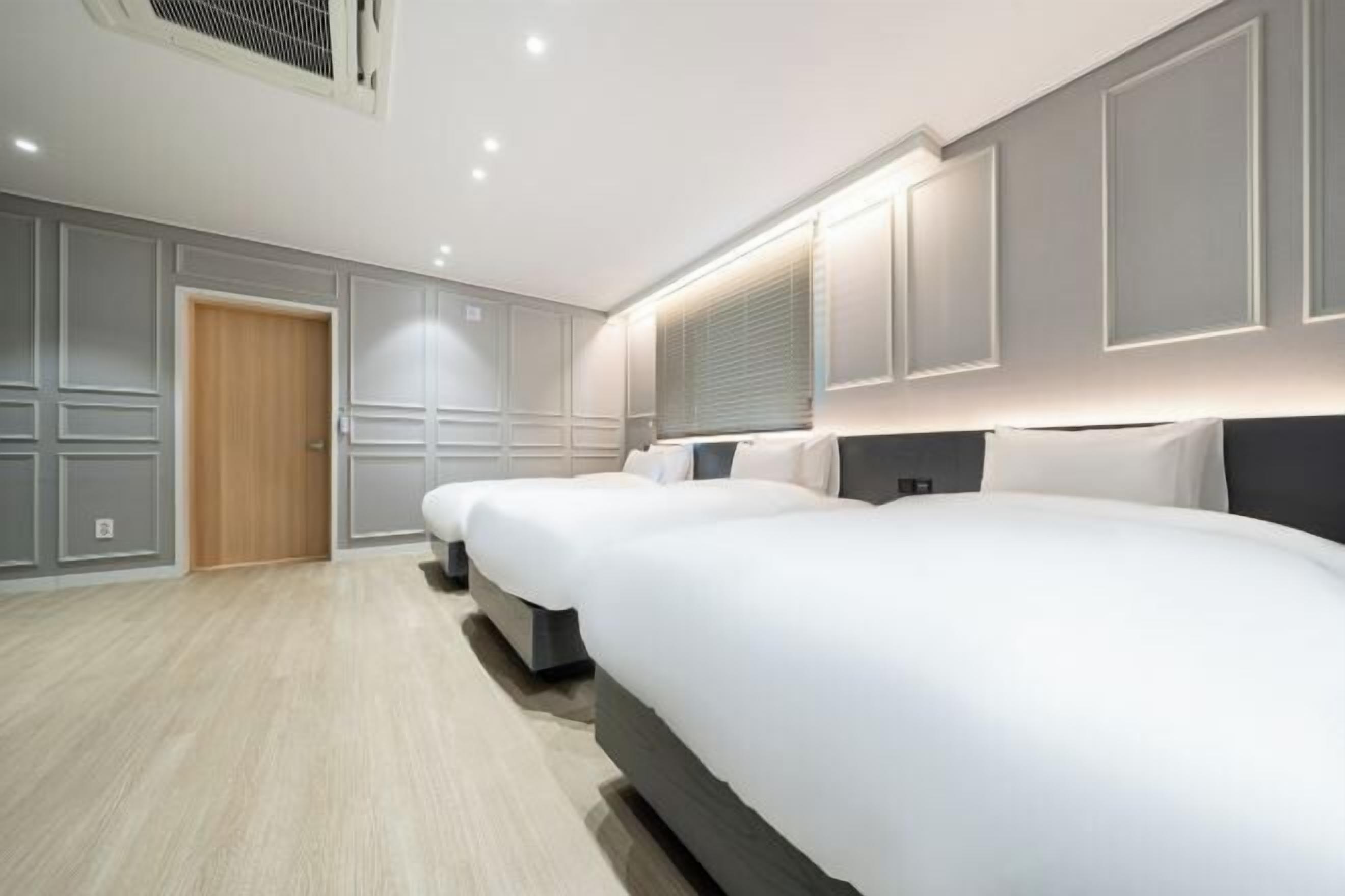 A hotel room with three beds aligned against the wall, a wooden door, and a window with blinds.