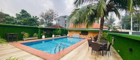 Une piscine dotée d’une terrasse en bois et de chaises, entourée de verdure et d’un palmier.