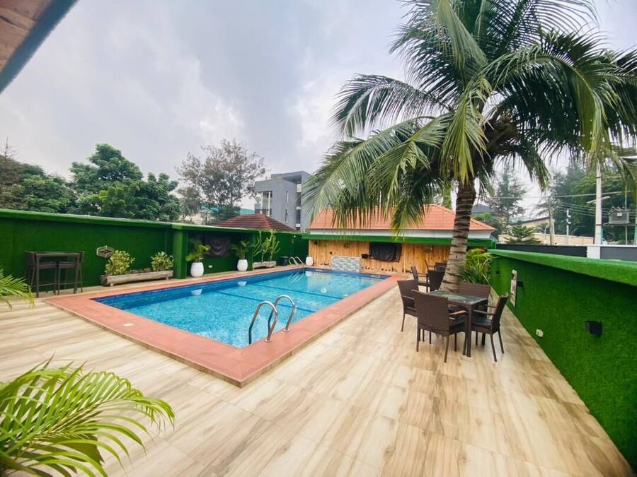 Una piscina con cubierta de madera y sillas, rodeada de follaje y una palmera.