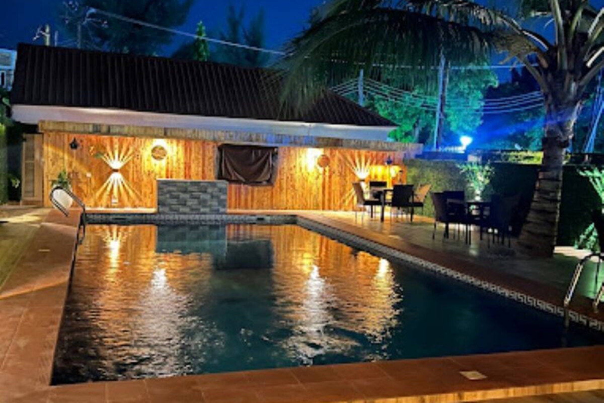 A swimming pool with a wooden deck, a small building with a tiled roof, and palm trees.