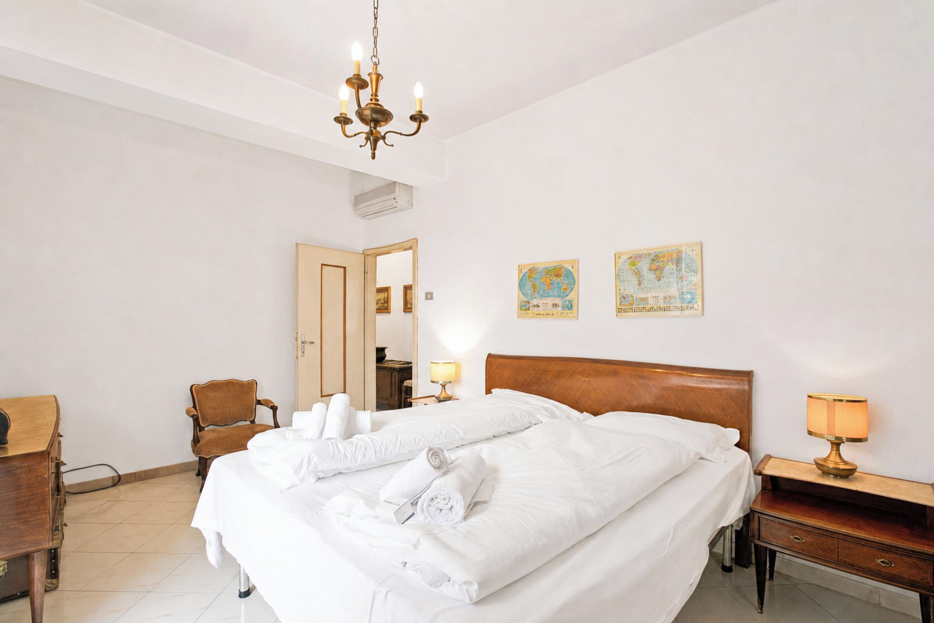 A hotel room with a large bed, a wooden headboard, a desk, a chair, a nightstand, a lamp, and two wall maps.