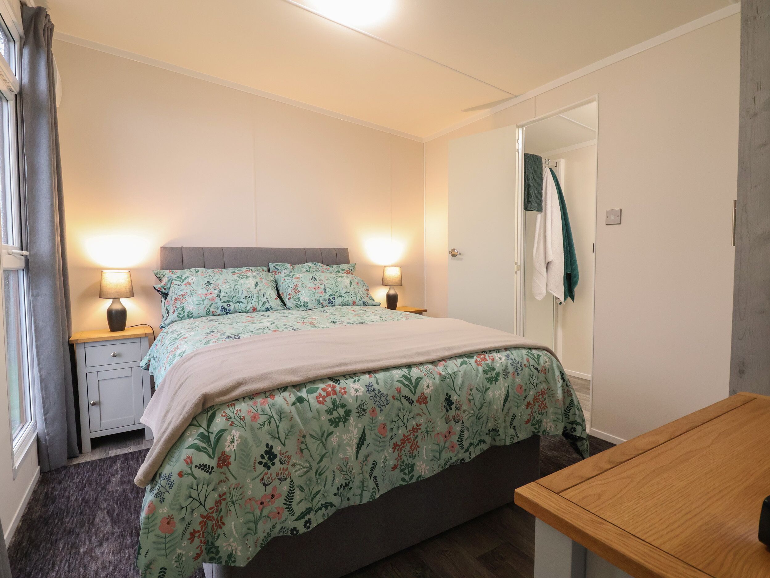 A bedroom with a bed, bedside lamps, a wooden table, and a bathroom door.