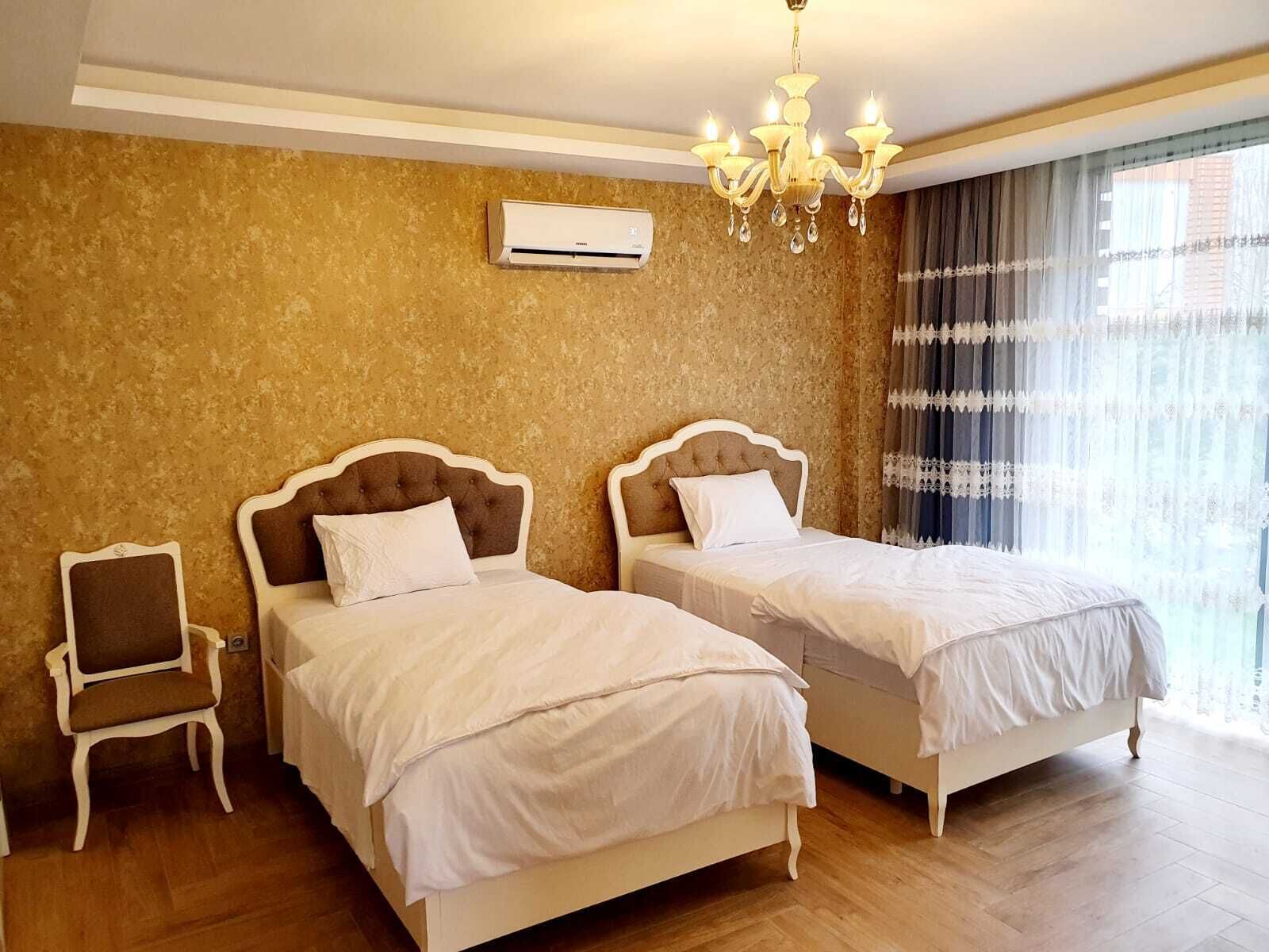 Habitación de hotel con dos camas, un candelabro, un equipo de aire acondicionado y vistas al exterior a través de cortinas translúcidas.