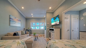 A cozy room with a sofa, a television mounted on the wall, and a window with blinds. - Family Studio • Beach Boardwalk + Shared Pools | Golf Cart Zone | Beach Bungalow (Port Aransas)