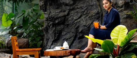 A woman sitting on a wooden chair at a table with a teapot and cup, outdoors with a rock wall and greenery in the background.
