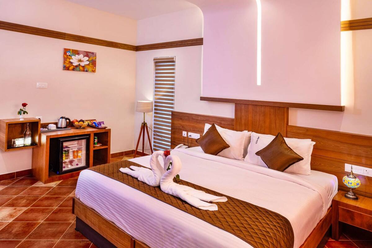 A hotel room with a large bed, a ceiling fan, a bedside table, a nightstand, a lamp, a small shelf with decorative items, and a window with blinds.