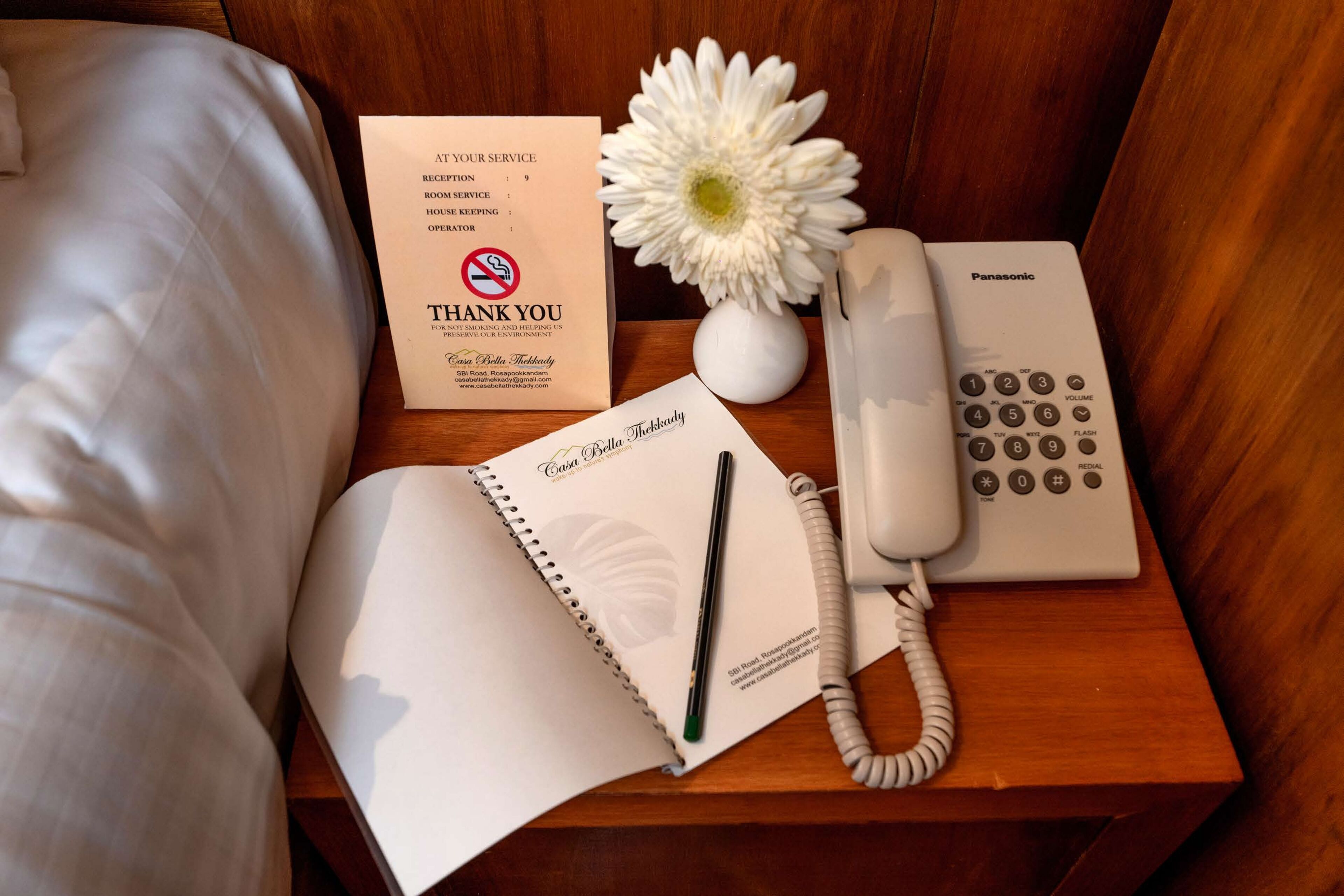 Een keurig opgemaakt hotelkamer met een bed, een nachtkastje met een witte bloevas, een notitieboekje, een pen, een Panasonic telefoon en een bedankkaart.