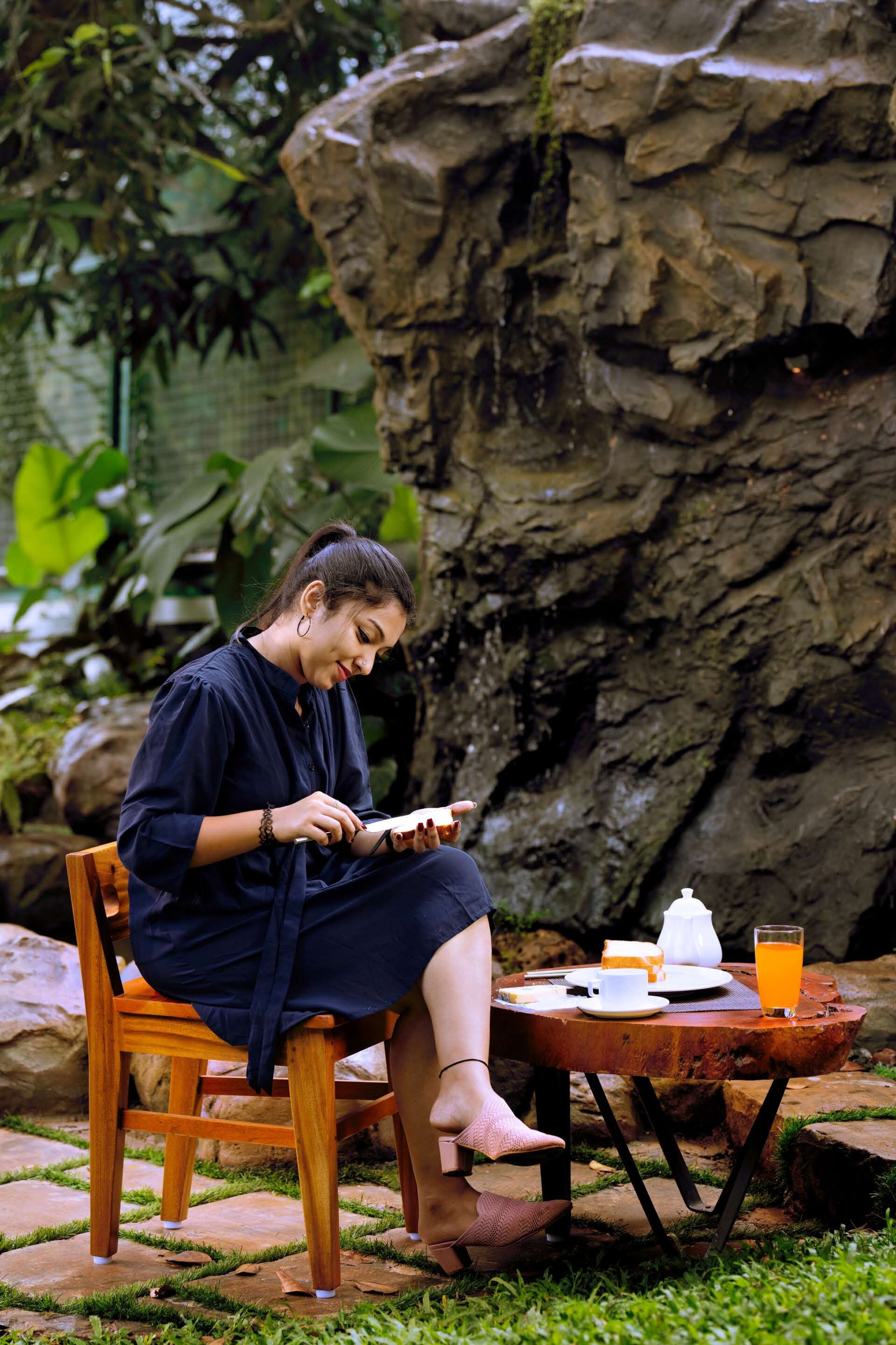 Une personne est assise sur une chaise en bois à l'extérieur, avec une table à côté d'elle sur laquelle se trouvent une théière, une tasse et une boisson à l'orange.