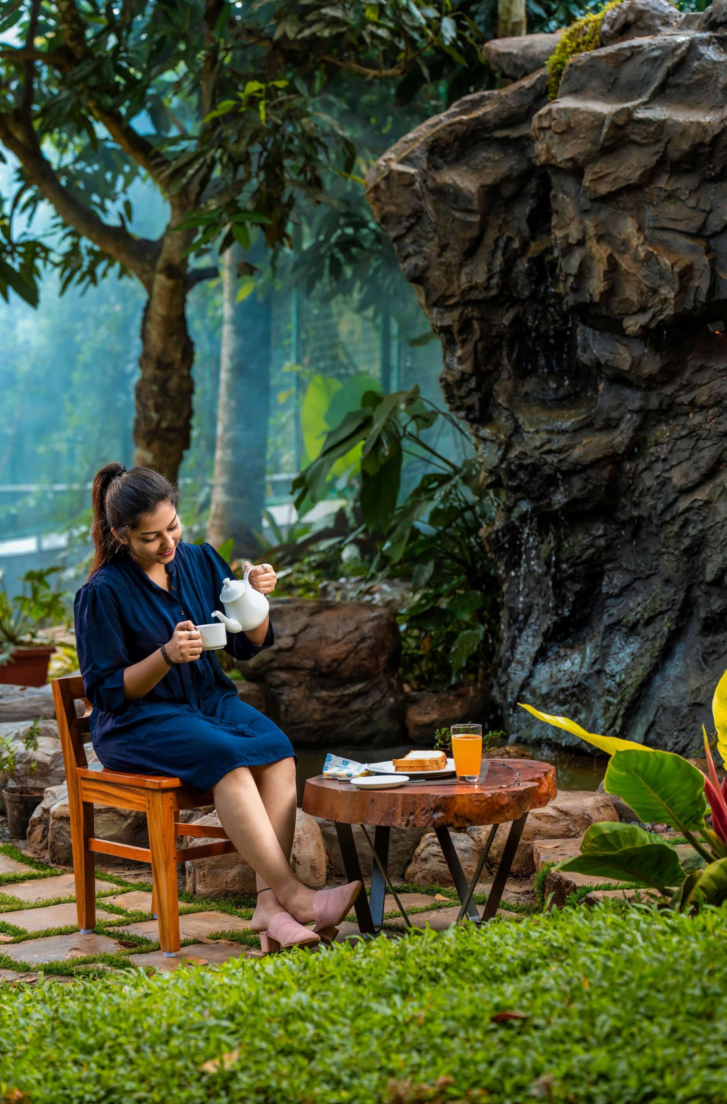 A person sitting on a wooden chair in a lush garden setting, holding a teapot and a cup.