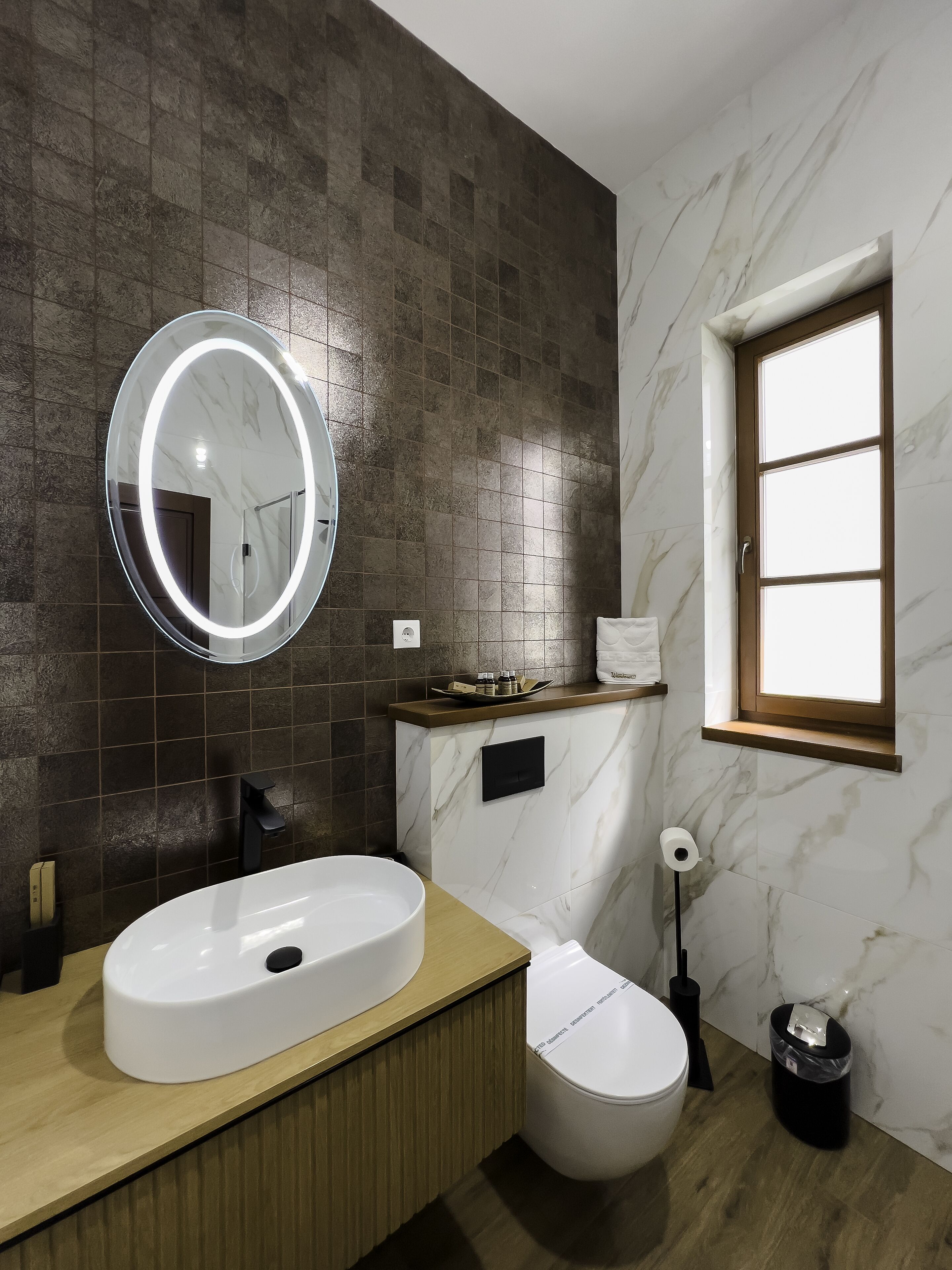 Ein modernes Badezimmer mit einem großen, ovalen Waschbecken, einem runden Spiegel mit Beleuchtung, einer Toilette und einem Fenster.