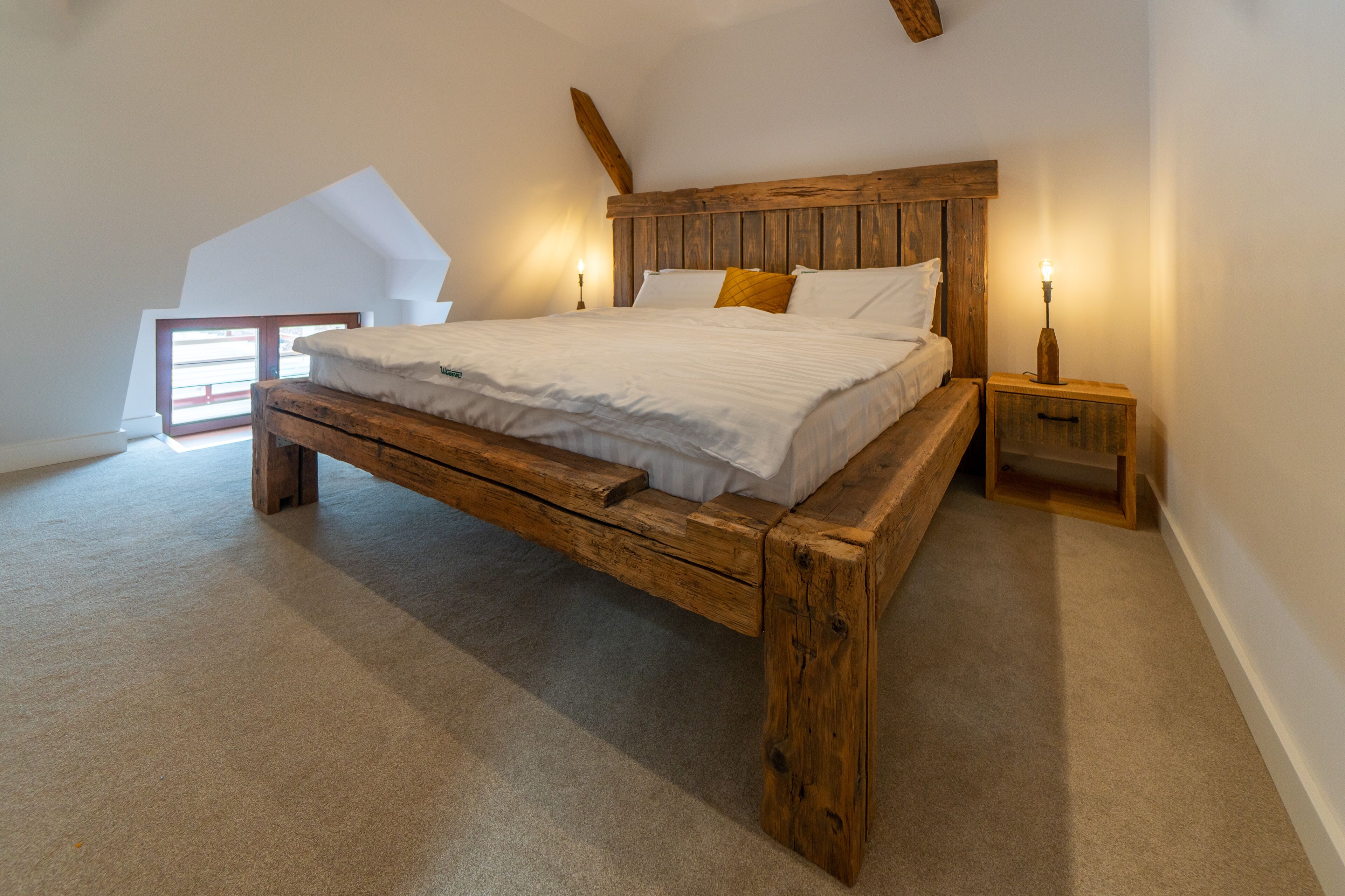 Quarto com estrutura de cama de madeira, roupa de cama branca e duas mesas de cabeceira com abajures.
