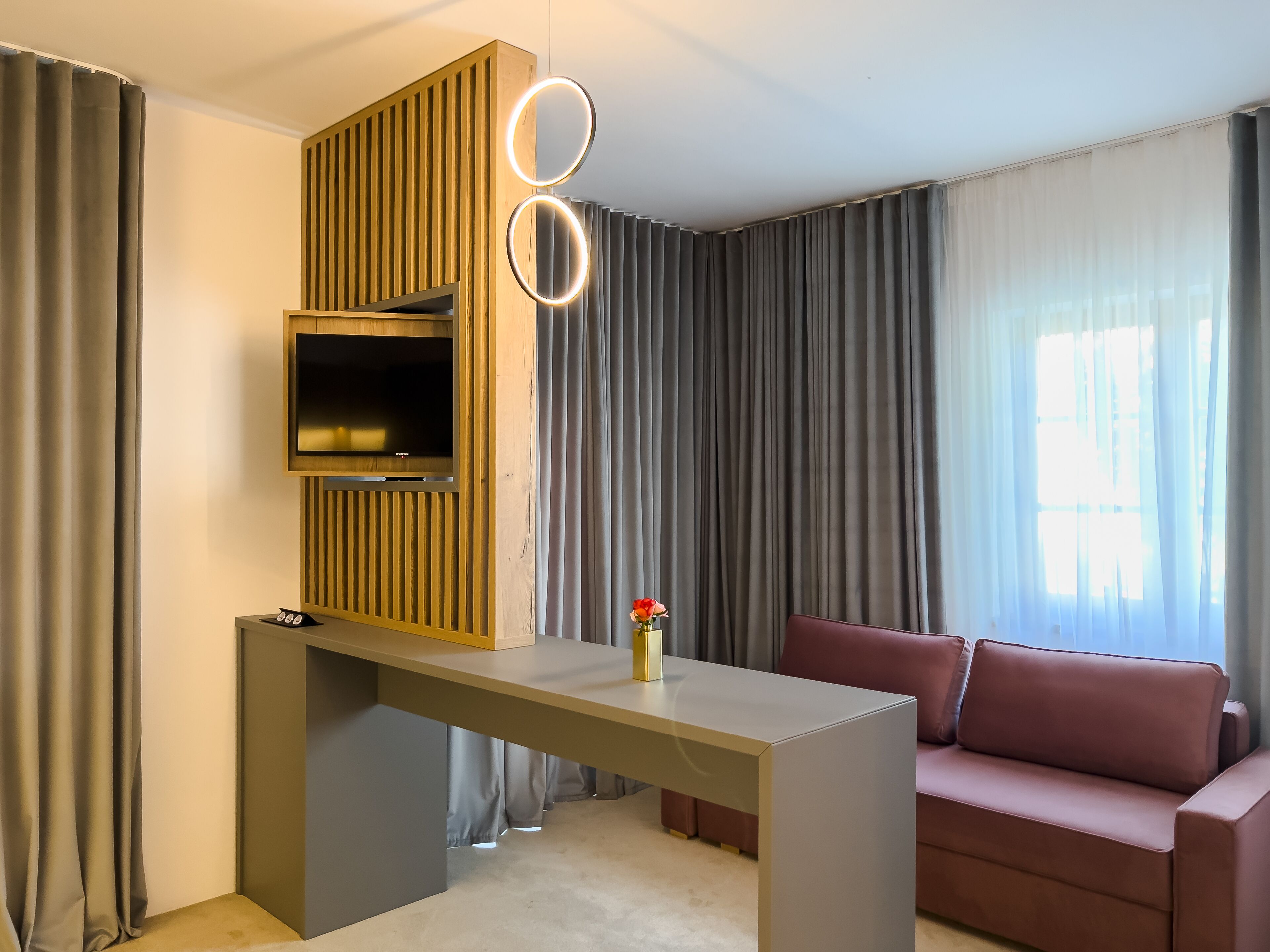 A modern living room with a TV mounted on a wooden panel wall, a desk with a ring light, a red sofa, and a window with curtains.
