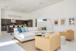 Un salon moderne avec un canapé blanc, des fauteuils beige, une table basse et un coin cuisine doté de meubles sombres et d’une hotte de cuisine en acier inoxydable.