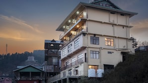 Un edificio de varios pisos con balcones y ventanas grandes, ubicado en una ladera con otros edificios al fondo.