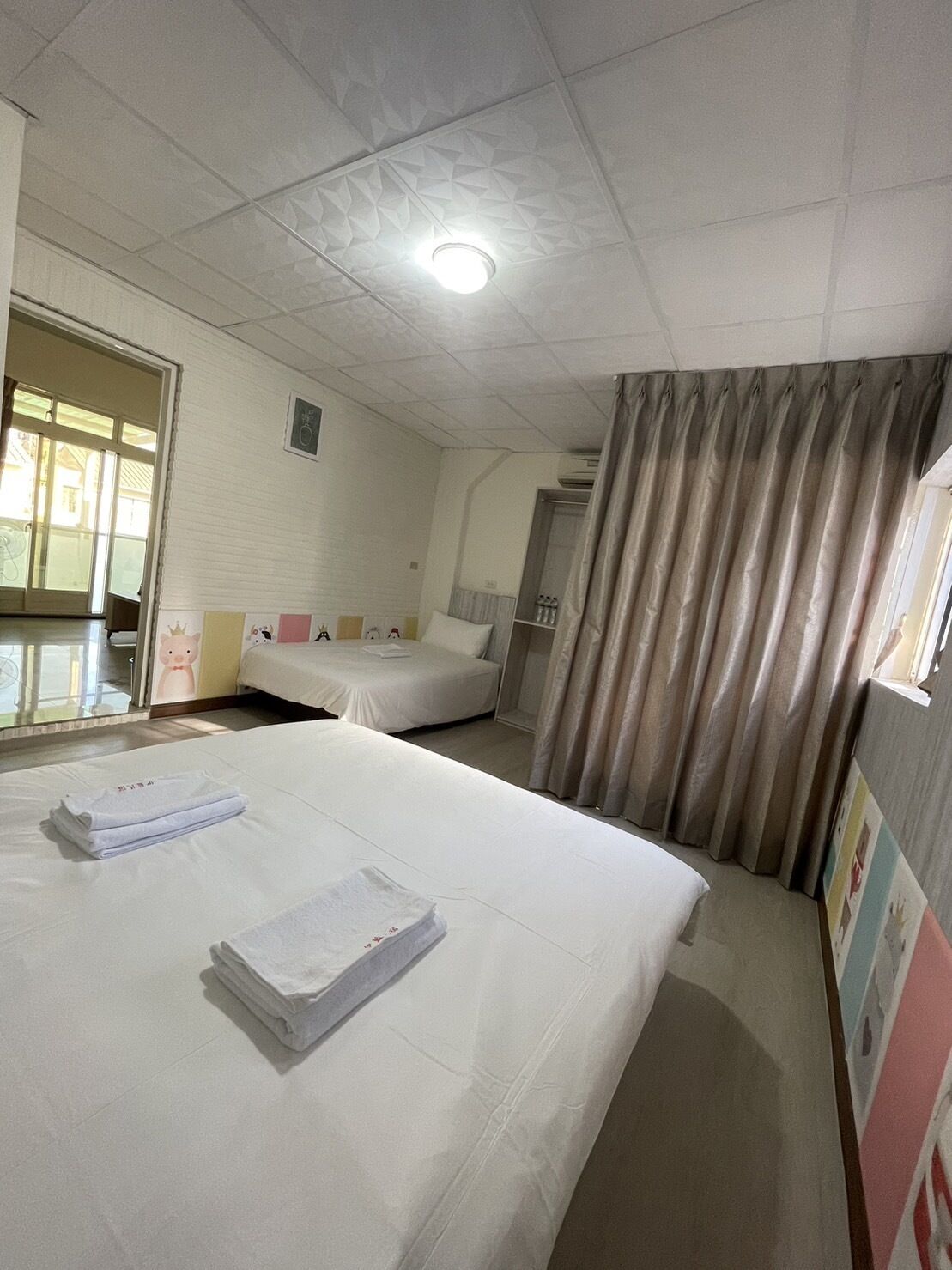 A hotel room with two beds, a large window, a curtain, and a ceiling light.