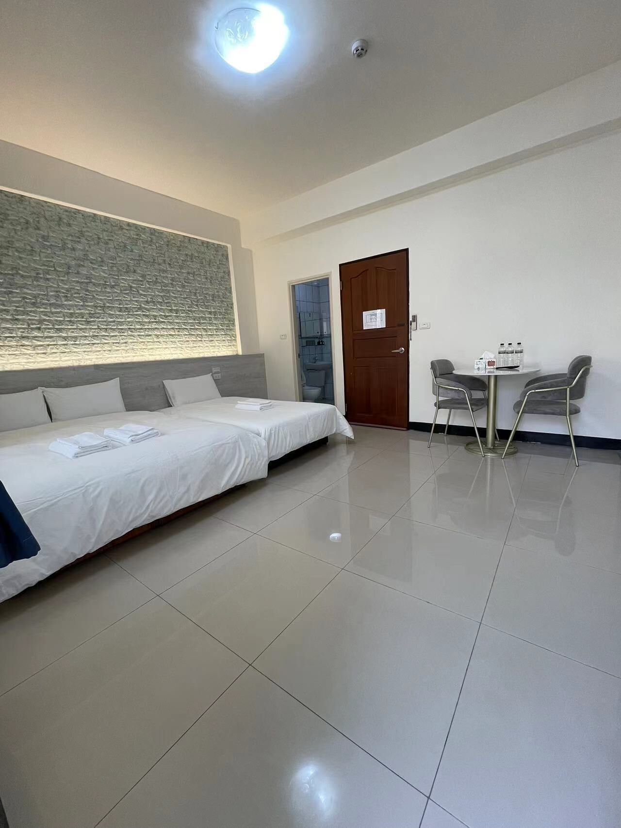 Habitación de hotel con una cama grande, una mesita con sillas, una puerta y un baño visible a través de una puerta abierta.