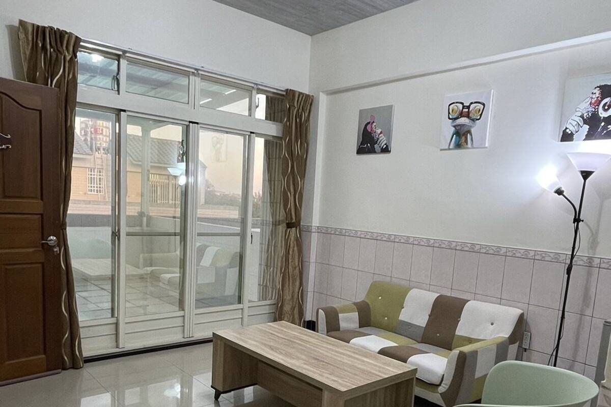 A spacious living room with a wooden coffee table, a sofa, and a light-colored armchair. A sliding glass door leads to an outdoor area.