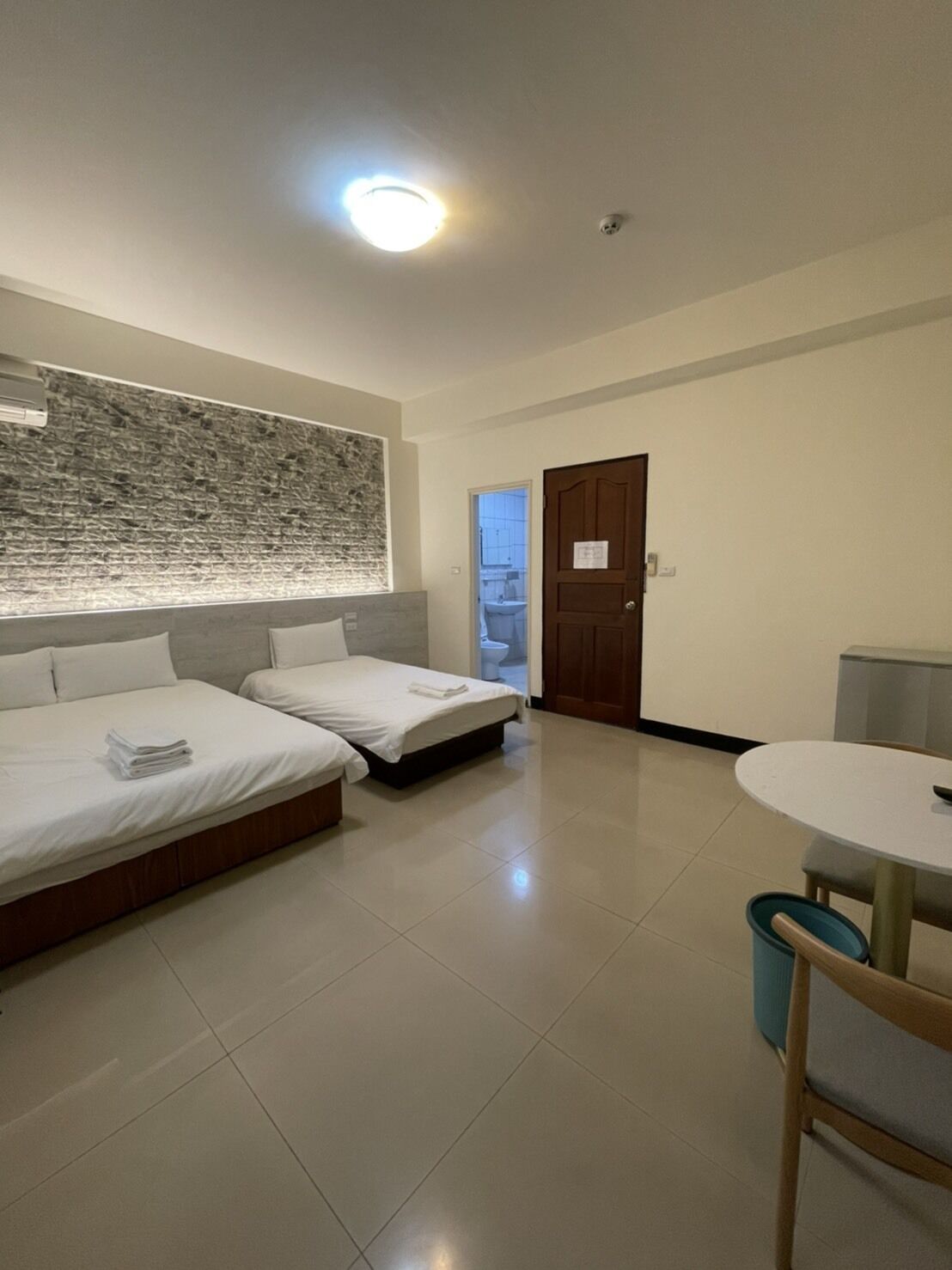 Habitación de hotel con dos camas, una mesita redonda pequeña, un aire acondicionado instalado en la pared y una puerta que da a un baño.
