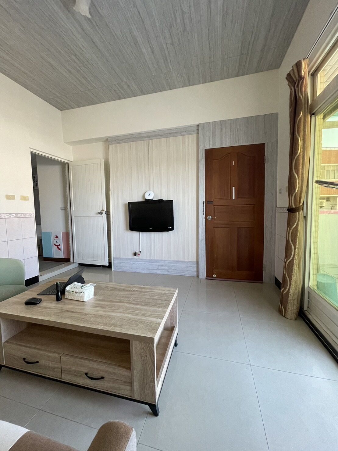 A modern living room with a wooden coffee table, a flat-screen TV mounted on the wall, a wooden door, and a large window with curtains.