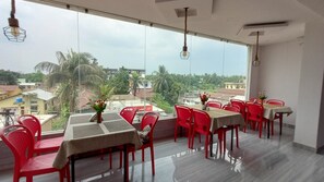 A dining area with red chairs and tables, large windows with a view of buildings and trees, and hanging light fixtures. - Roy's Hotel (Agomoni)