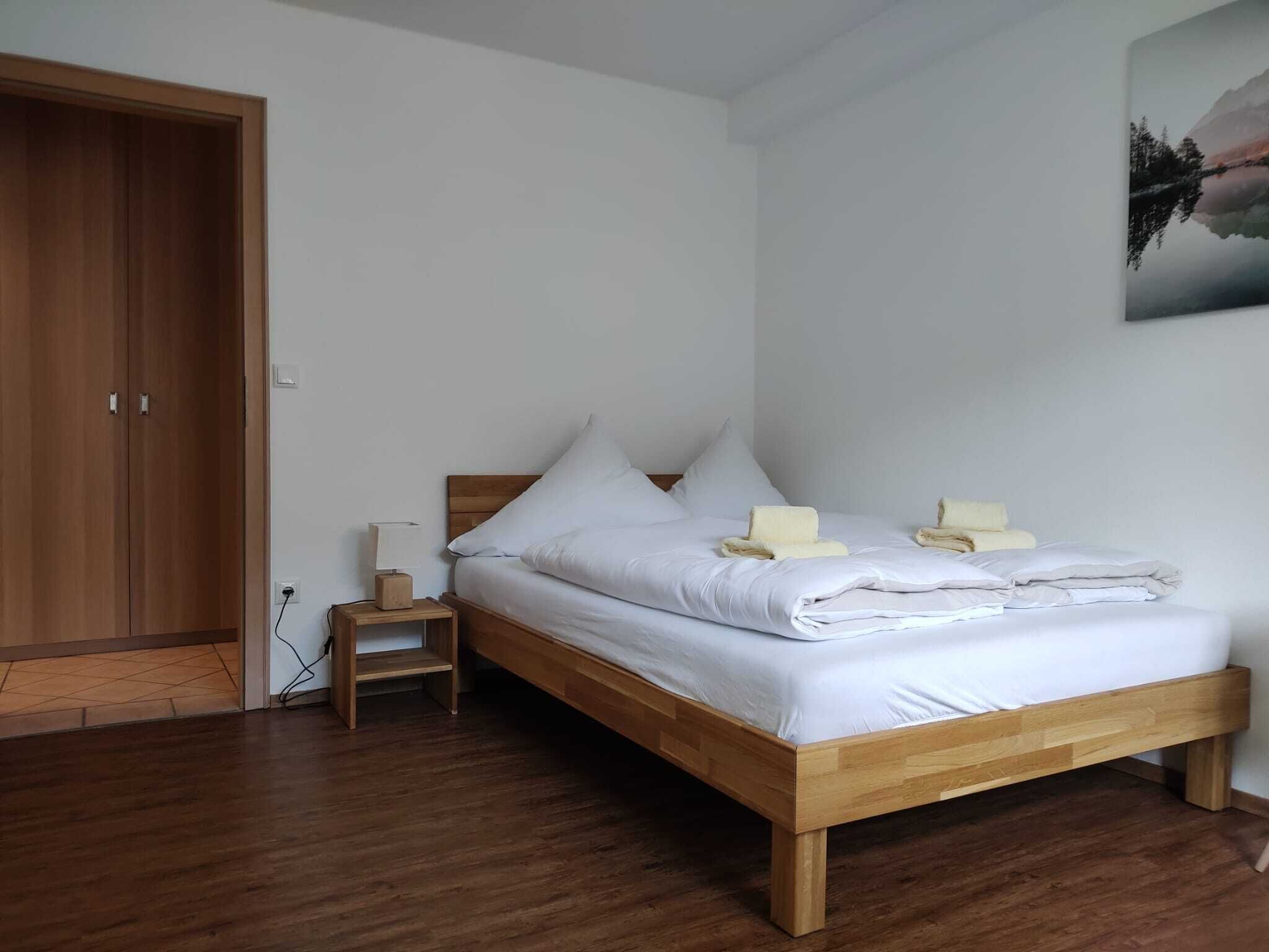 A neatly made bed with white linens and pillows, a wooden nightstand with a lamp, and a wooden wardrobe.