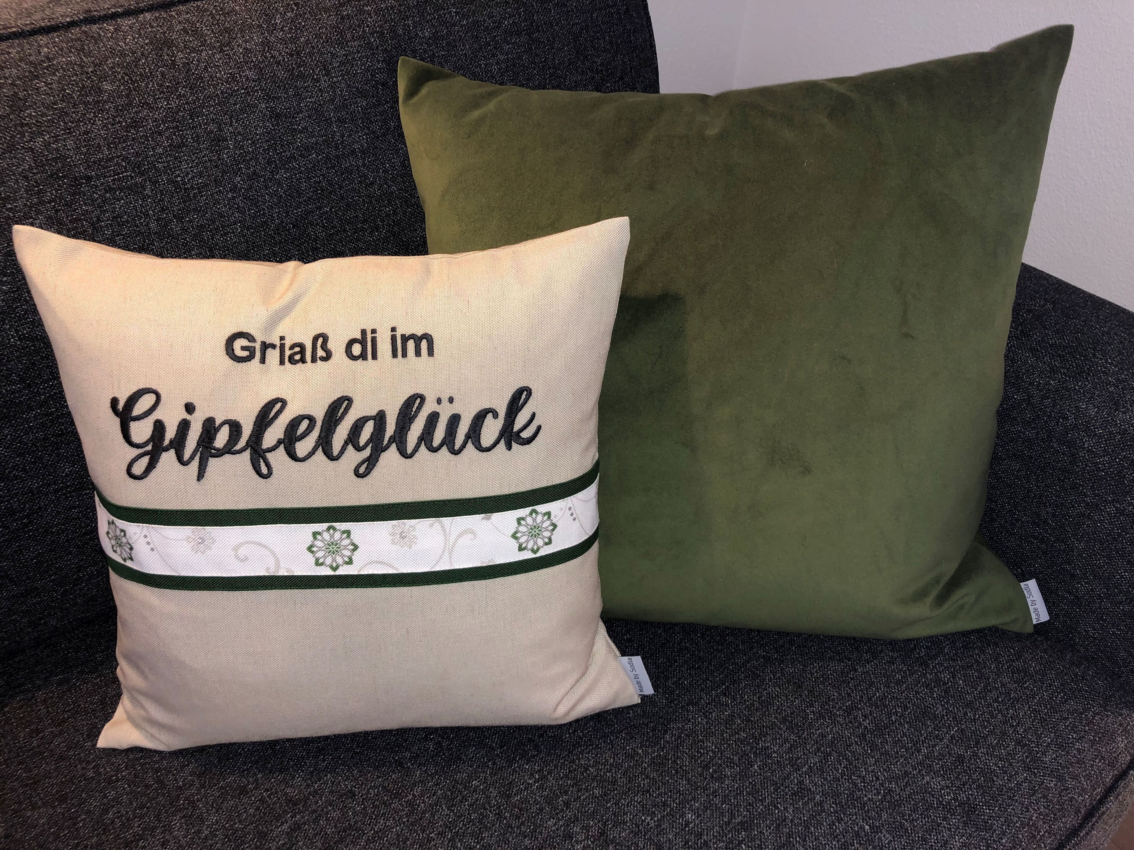 A beige pillow with embroidered text and a green ribbon, placed on a dark sofa.