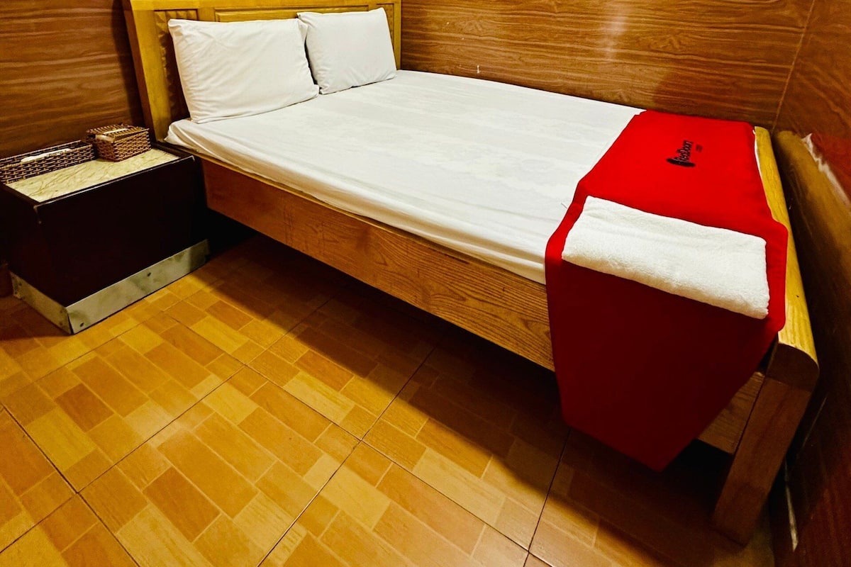 A single bed with a wooden headboard and footboard, a white mattress, and a red blanket. There is a small wooden nightstand with a basket beside the bed. The floor is tiled with a wooden pattern.