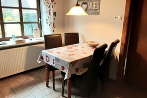 Un comedor con una mesa cubierta por un mantel estampado, dos sillas, un bol en la mesa y una ventana con cortinas.