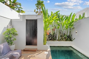 A modern outdoor pool area with a wooden deck, a built-in bench, and a variety of tropical plants. - Liminal Palace Villa 3 by Betterplace (Canggu)