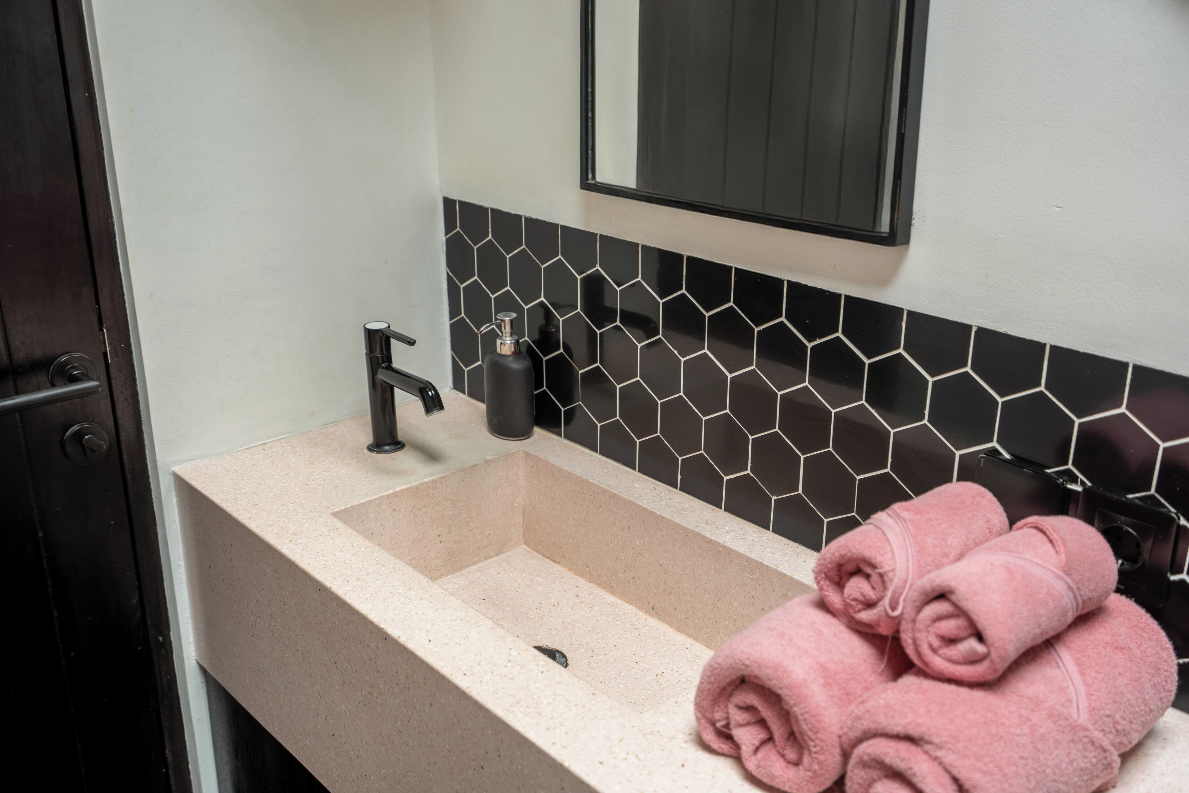A bathroom with a beige sink, black hexagonal tiles, a mirror, and two pink towels.