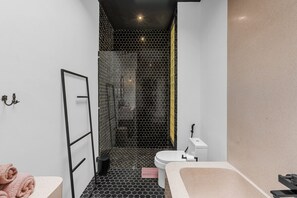 A modern bathroom with a black and white hexagonal tile floor, a white toilet, a beige bathtub, and a glass shower enclosure. - Liminal Palace Villa 3 by Betterplace (Canggu)