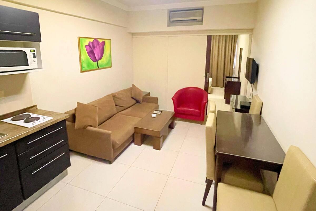 A modern living room with a sofa, coffee table, and dining area. There is a kitchen area with a microwave and a TV.