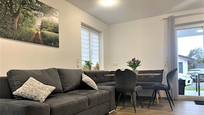 Un salon moderne avec un canapé d’angle gris foncé, une table à manger avec des chaises, une grande fenêtre avec des stores et une vue sur une voiture garée à l’extérieur.