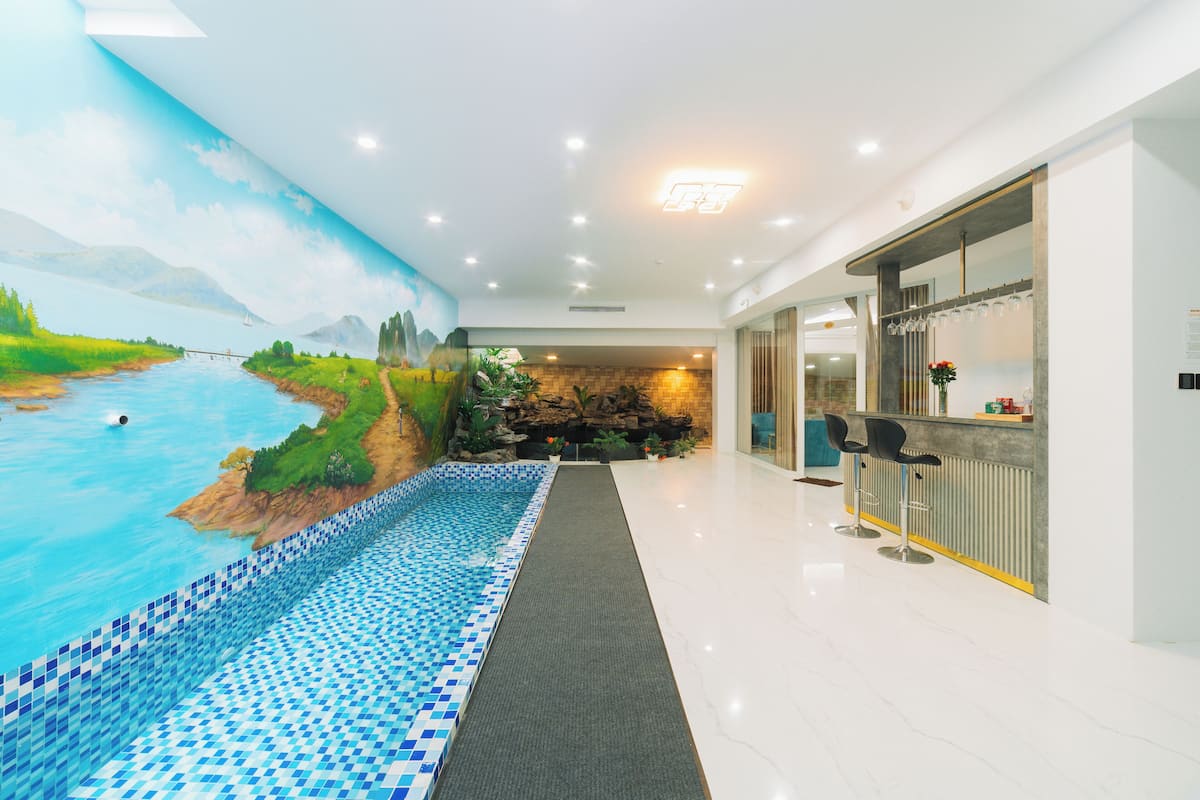 Un moderno vestíbulo de hotel con un gran mural de un paisaje fluvial, una zona de piscina con baldosas azules y blancas, y un bar con taburetes.
