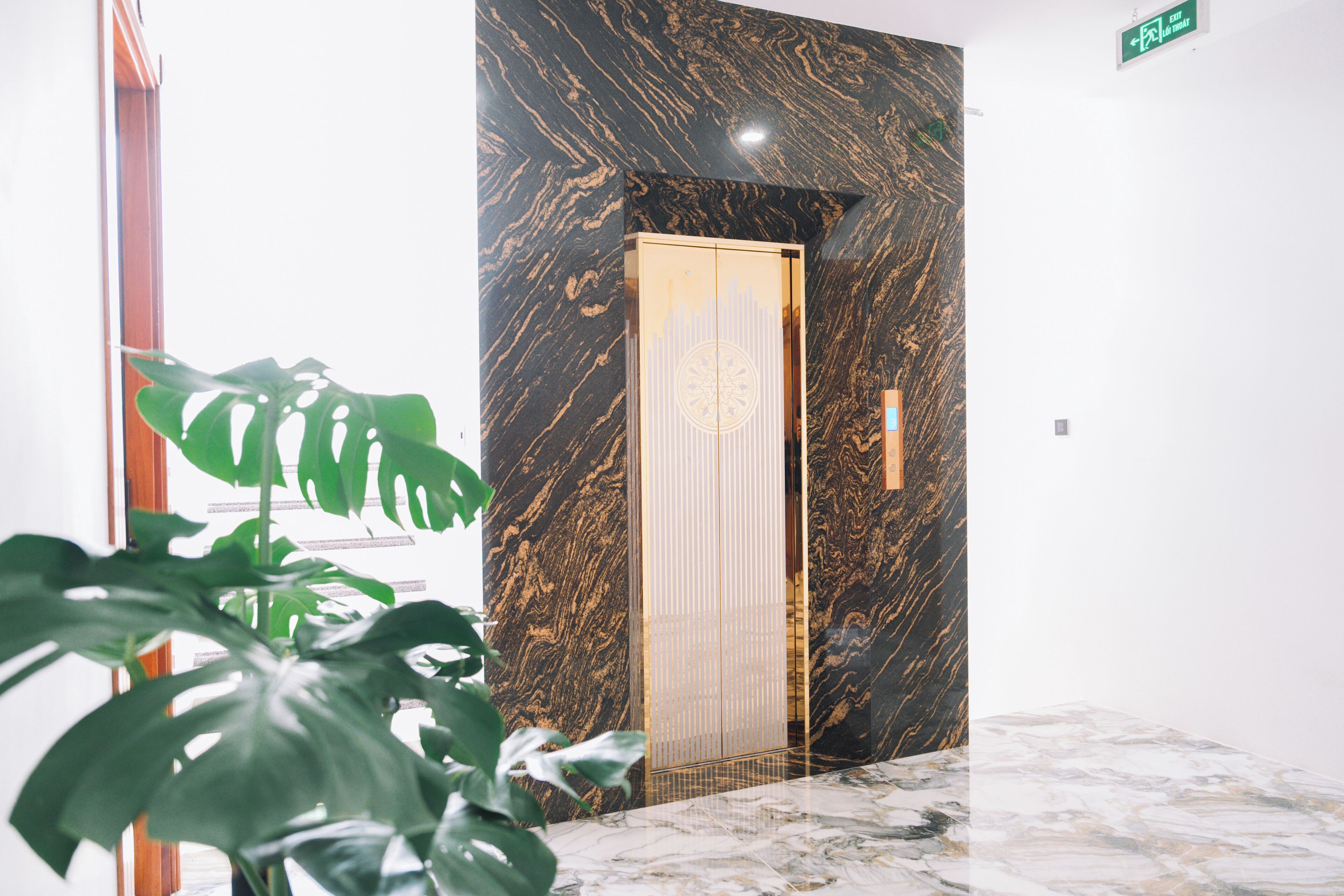 A modern elevator with a marble wall and a gold door, surrounded by a potted plant.